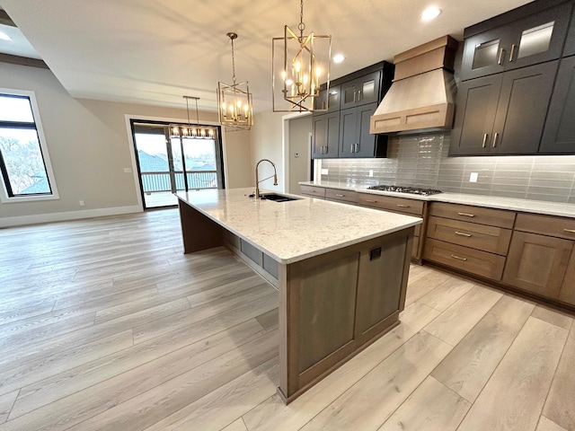 kitchen featuring light stone counters, custom exhaust hood, a kitchen island with sink, pendant lighting, and stainless steel gas stovetop