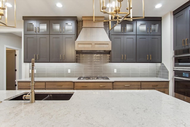 kitchen featuring stainless steel gas cooktop, custom range hood, backsplash, light stone counters, and glass insert cabinets
