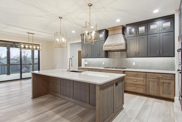 kitchen featuring a chandelier, a kitchen island with sink, a sink, light wood-type flooring, and custom range hood