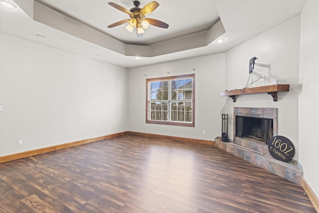 unfurnished living room with a tray ceiling, a tiled fireplace, ceiling fan, and dark wood-type flooring