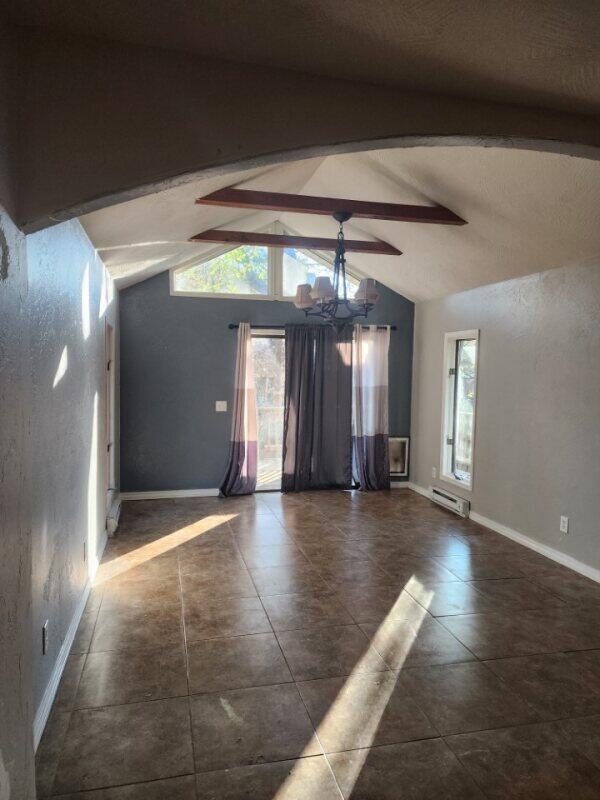 tiled empty room with a baseboard heating unit, lofted ceiling with beams, a wealth of natural light, and ceiling fan with notable chandelier