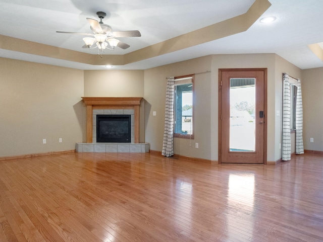 unfurnished living room featuring a tiled fireplace, light hardwood / wood-style floors, ceiling fan, and a tray ceiling