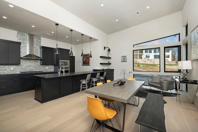 kitchen with a center island with sink, wall chimney range hood, hanging light fixtures, light hardwood / wood-style floors, and stainless steel appliances