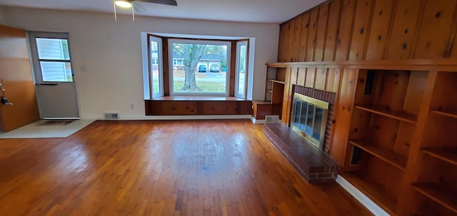 unfurnished living room featuring hardwood / wood-style flooring, a brick fireplace, ceiling fan, and wooden walls