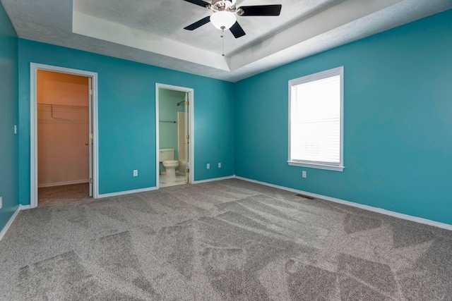 unfurnished bedroom featuring carpet, a raised ceiling, a walk in closet, ensuite bath, and ceiling fan