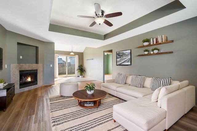 living room featuring a raised ceiling, a tile fireplace, ceiling fan, and wood-type flooring