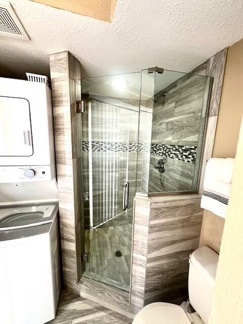 bathroom featuring walk in shower, toilet, a textured ceiling, and stacked washing maching and dryer