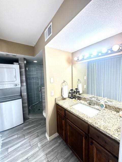 bathroom with a shower with door, stacked washing maching and dryer, vanity, a textured ceiling, and toilet