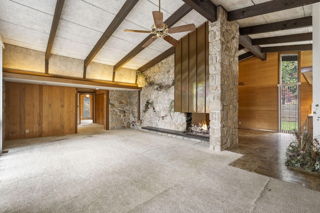 unfurnished living room featuring vaulted ceiling with beams, ceiling fan, a stone fireplace, and wooden walls