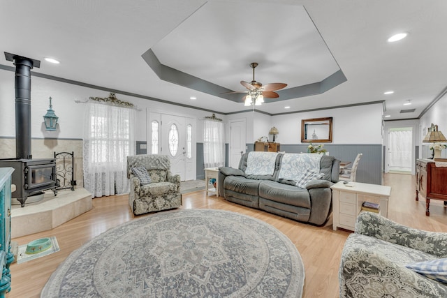 living room featuring a wood stove, ceiling fan, light wood-type flooring, a tray ceiling, and ornamental molding