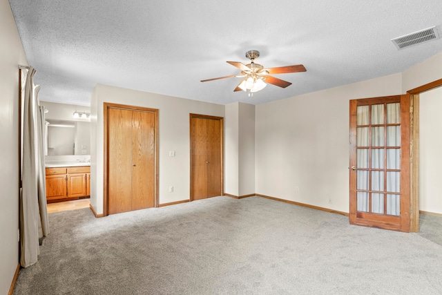 unfurnished bedroom featuring ensuite bath, a textured ceiling, light colored carpet, ceiling fan, and multiple closets