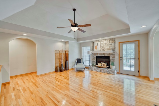 unfurnished room with ceiling fan, a stone fireplace, a tray ceiling, and light hardwood / wood-style flooring