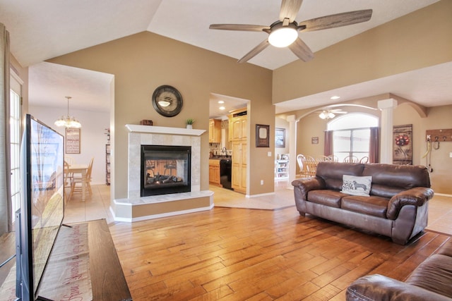 living room with ceiling fan with notable chandelier, a tiled fireplace, vaulted ceiling, and light wood-type flooring