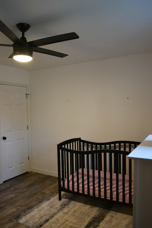 bedroom with a crib, ceiling fan, and dark wood-type flooring