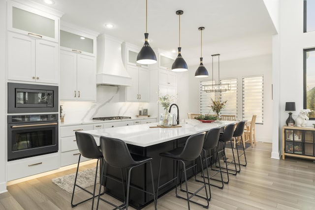 kitchen with white cabinetry, hanging light fixtures, a kitchen island with sink, black appliances, and custom exhaust hood