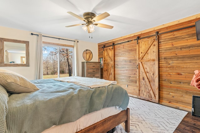bedroom featuring access to exterior, ceiling fan, wooden walls, a barn door, and dark hardwood / wood-style floors