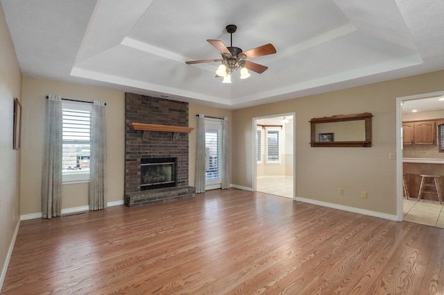 unfurnished living room with a brick fireplace, ceiling fan, light wood-type flooring, and a tray ceiling