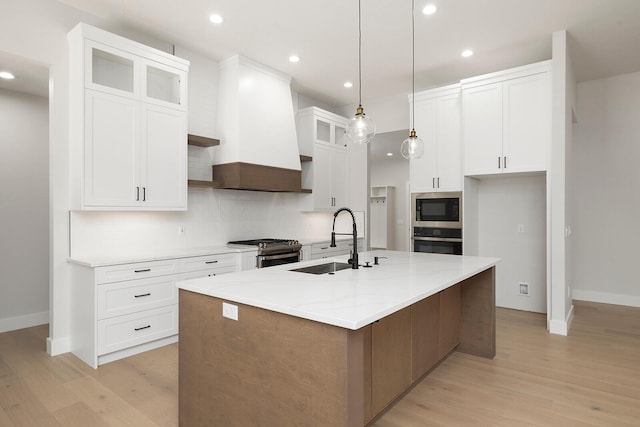 kitchen featuring premium range hood, a kitchen island with sink, white cabinets, light stone counters, and stainless steel appliances