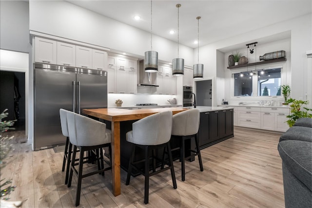 kitchen featuring pendant lighting, white cabinets, wall chimney exhaust hood, an island with sink, and stainless steel appliances