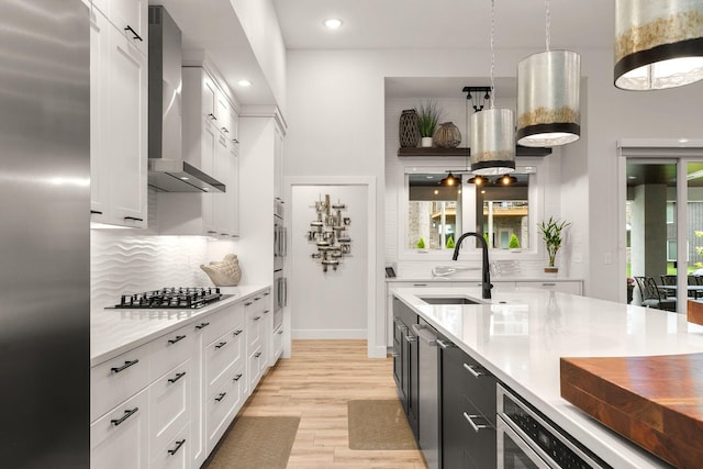 kitchen with sink, stainless steel appliances, wall chimney range hood, pendant lighting, and white cabinets