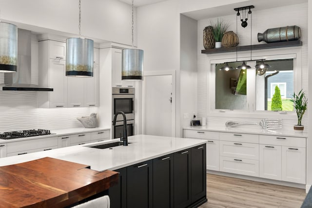 kitchen with white cabinets, hanging light fixtures, wall chimney exhaust hood, light wood-type flooring, and stainless steel appliances