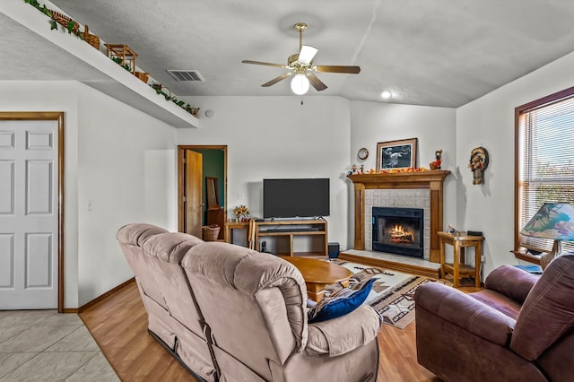 living room with ceiling fan, light hardwood / wood-style floors, a textured ceiling, vaulted ceiling, and a tiled fireplace