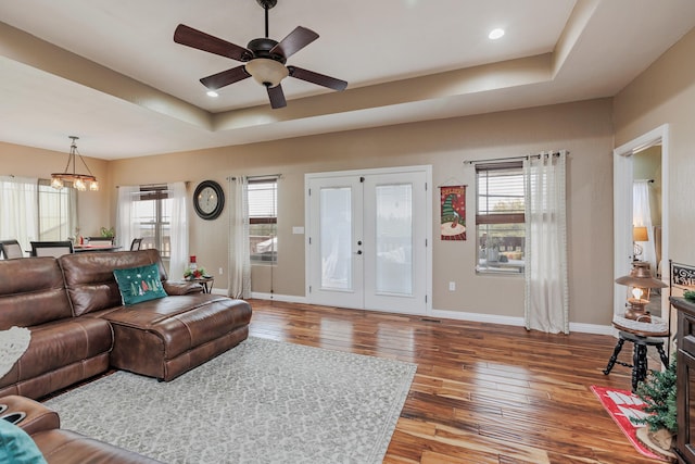 living room with baseboards, hardwood / wood-style flooring, a tray ceiling, french doors, and ceiling fan with notable chandelier