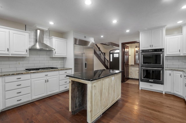 kitchen featuring decorative backsplash, appliances with stainless steel finishes, dark wood-type flooring, wall chimney range hood, and dark stone counters