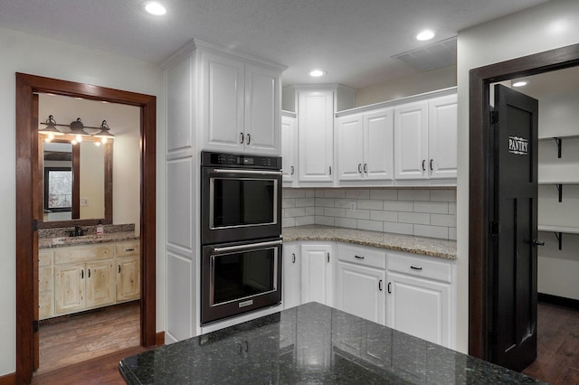 kitchen with dobule oven black, dark wood-style flooring, white cabinets, dark stone counters, and tasteful backsplash