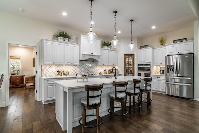 kitchen featuring dark wood-type flooring, white cabinets, a center island with sink, decorative light fixtures, and stainless steel appliances