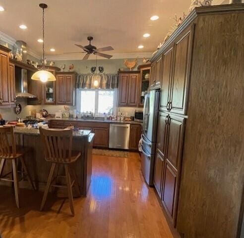 kitchen featuring wall chimney exhaust hood, stainless steel appliances, ceiling fan, dark wood-type flooring, and a breakfast bar area