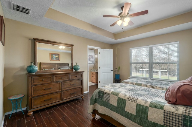 bedroom with ensuite bath, a textured ceiling, a tray ceiling, ceiling fan, and dark wood-type flooring