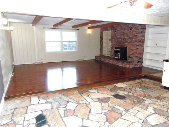 unfurnished living room featuring beam ceiling, a wood stove, ceiling fan, and wood-type flooring