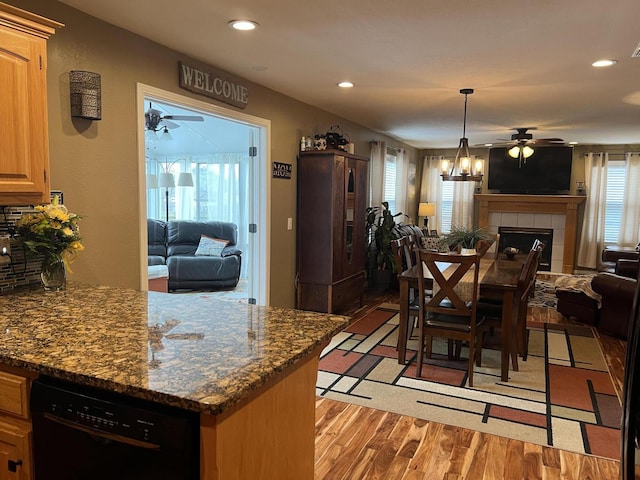 kitchen featuring ceiling fan, black dishwasher, dark stone countertops, a tiled fireplace, and light wood-type flooring