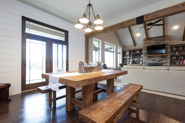 dining area featuring ceiling fan with notable chandelier, a stone fireplace, wooden walls, vaulted ceiling with beams, and dark hardwood / wood-style floors