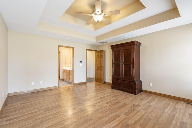 unfurnished bedroom featuring a raised ceiling, ceiling fan, ensuite bathroom, and light hardwood / wood-style floors