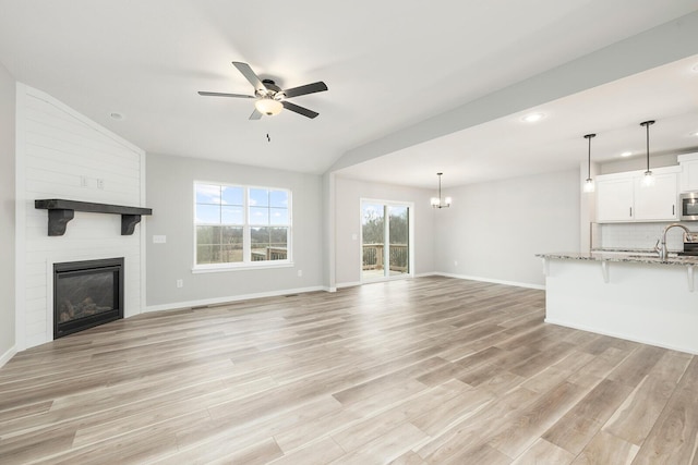 unfurnished living room with sink, a large fireplace, lofted ceiling, ceiling fan with notable chandelier, and light wood-type flooring