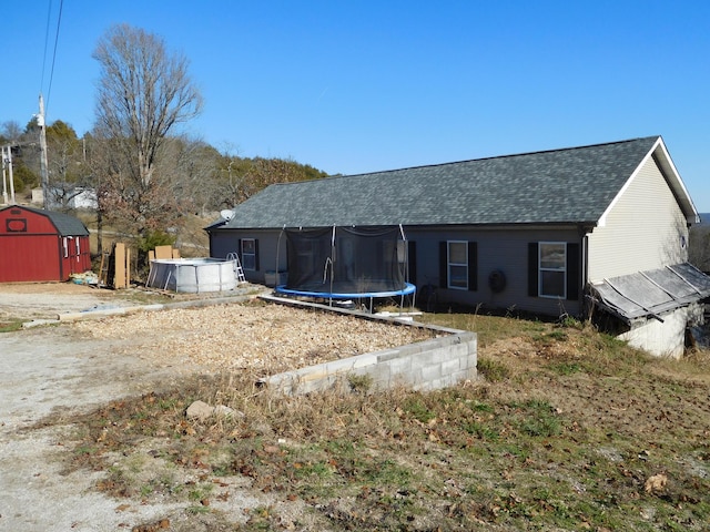 back of house with a trampoline, roof with shingles, an outdoor structure, and a shed