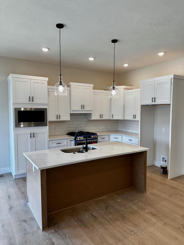 kitchen featuring decorative backsplash, a center island with sink, white cabinets, stainless steel microwave, and light wood finished floors