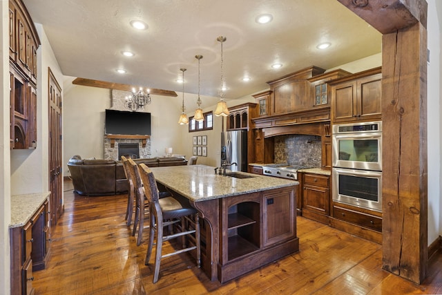 kitchen featuring a center island with sink, a stone fireplace, decorative light fixtures, light stone counters, and stainless steel appliances