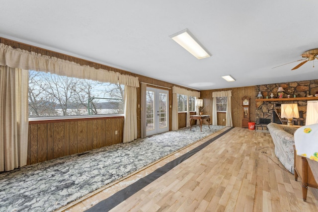 living area with french doors, ceiling fan, wood finished floors, a stone fireplace, and wood walls