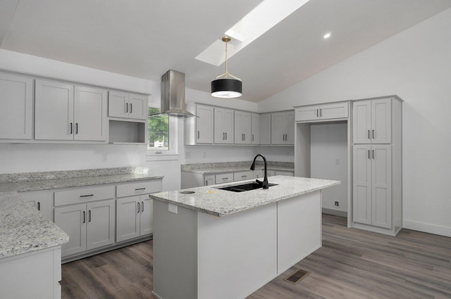 kitchen with light stone countertops, wall chimney exhaust hood, sink, a center island with sink, and hanging light fixtures