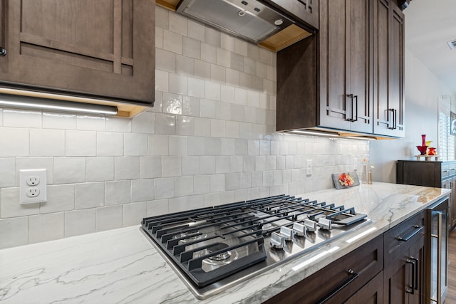 kitchen with decorative backsplash, light stone counters, dark brown cabinets, ventilation hood, and stainless steel gas cooktop