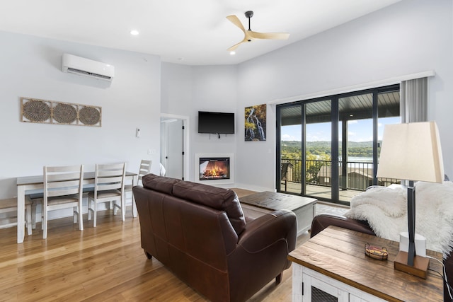 living room with a ceiling fan, a glass covered fireplace, a towering ceiling, a wall mounted air conditioner, and light wood-style floors