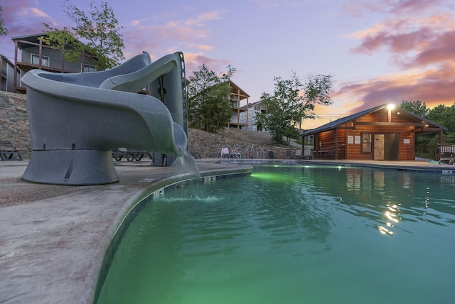 pool at dusk with an outdoor pool, a water slide, an exterior structure, a patio area, and an outdoor structure
