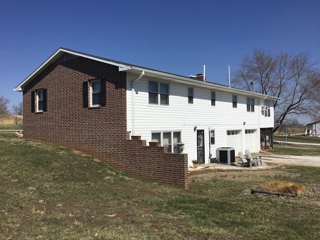 back of house with brick siding, an attached garage, central air condition unit, a lawn, and driveway