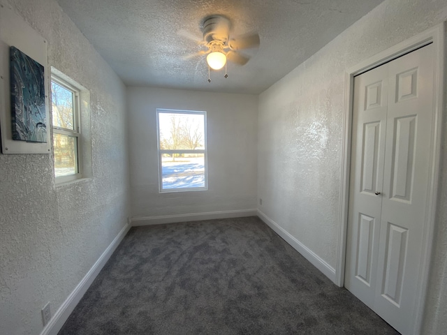 spare room featuring a textured ceiling, dark carpet, ceiling fan, and a healthy amount of sunlight