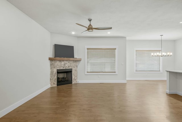 unfurnished living room with ceiling fan with notable chandelier, wood-type flooring, and a stone fireplace