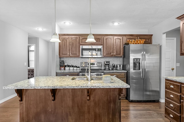 kitchen with stainless steel appliances, an island with sink, hanging light fixtures, light stone countertops, and dark hardwood / wood-style flooring