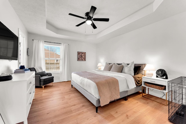 bedroom featuring ceiling fan, a textured ceiling, a tray ceiling, and light wood-type flooring